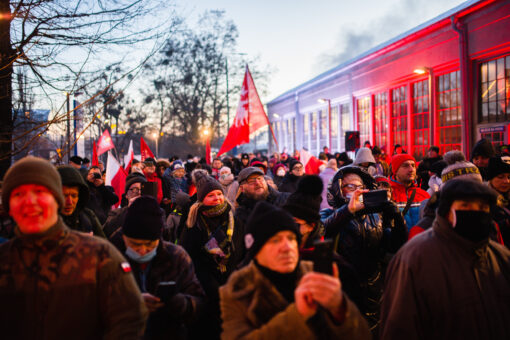 Tłum pod dworcem Letnim w Poznaniu. Wszycy brani zimowo. Ponad głowami łopoczą flagi powstańcze. W tle budynek dworca podświetlony na biało-czerwono.