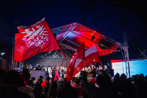 Tłum osób witających Ignacego Jana Paderwskiego w trakcie inscenizajcji jego przyjazdu do Poznania. Ponad głowami publiczności powiewają flagi powstańcze.